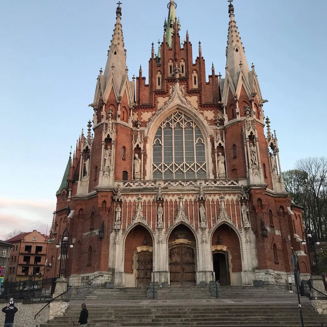 The image shows a large Gothic-style church made of red brick with ornate white stone details, tall spires, arched windows, and wooden doors at the entrance. Stepped stairs lead up to the doors, and there are a few people standing nearby.