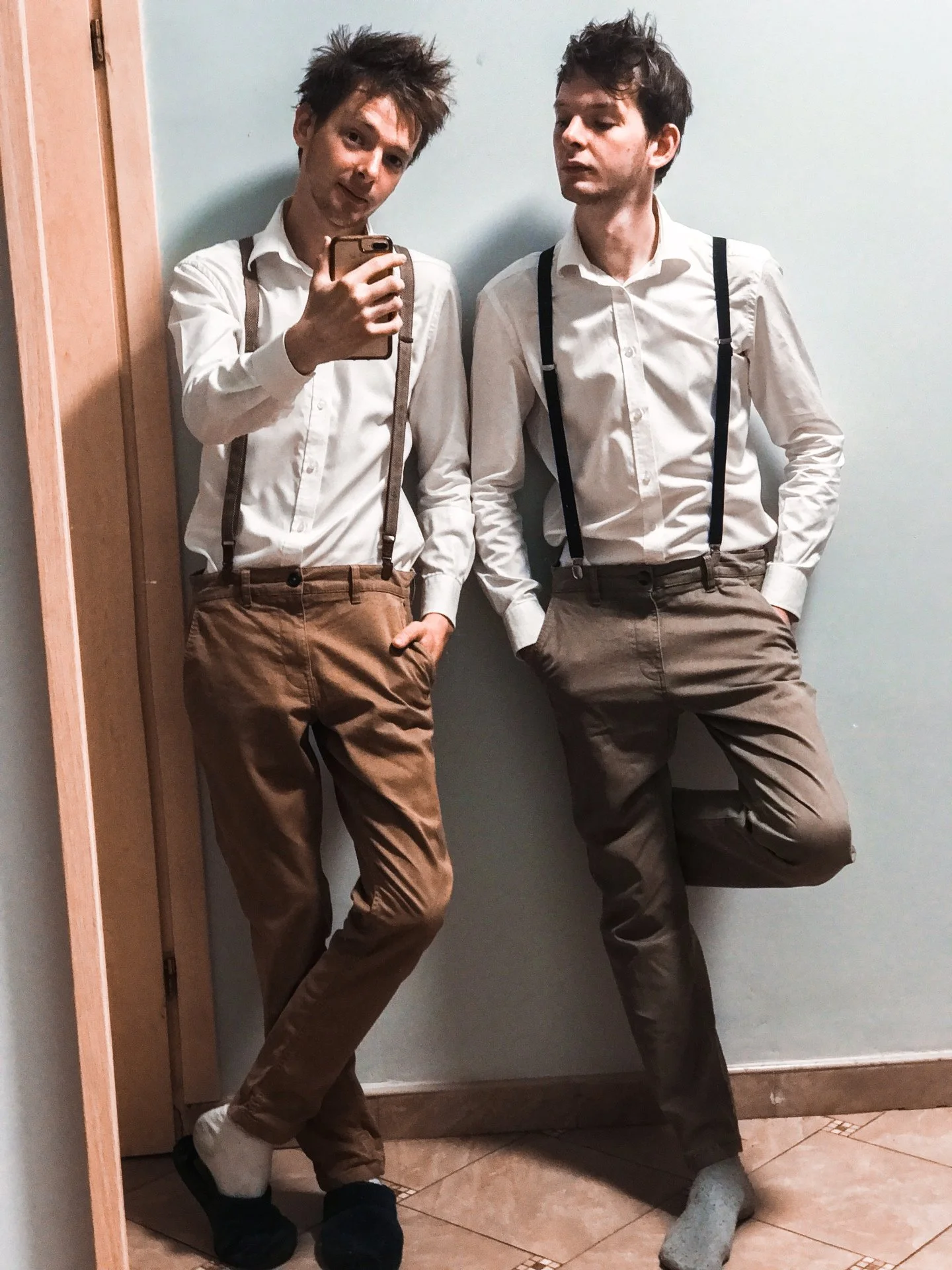 Two young men taking a mirror selfie, dressed in white shirts, suspenders, and brown pants, standing against a plain wall in a room.