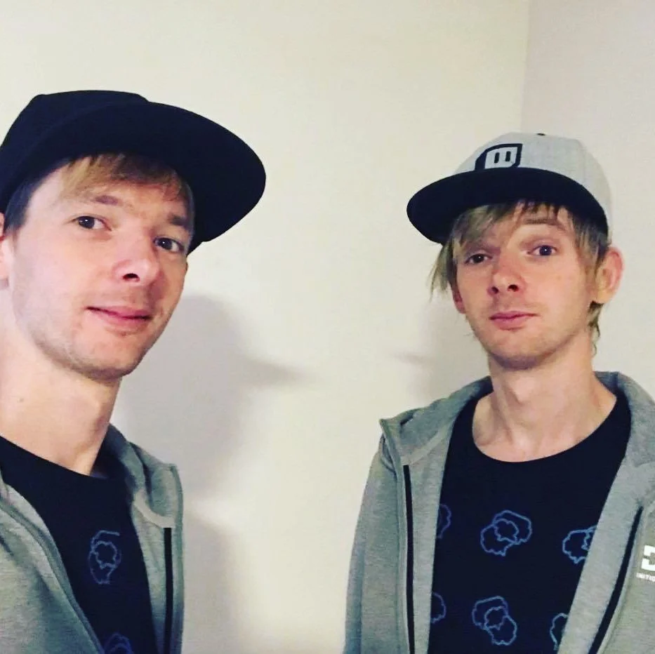 Two young men wearing baseball caps and gray jackets, standing against a plain wall, taking a selfie together.