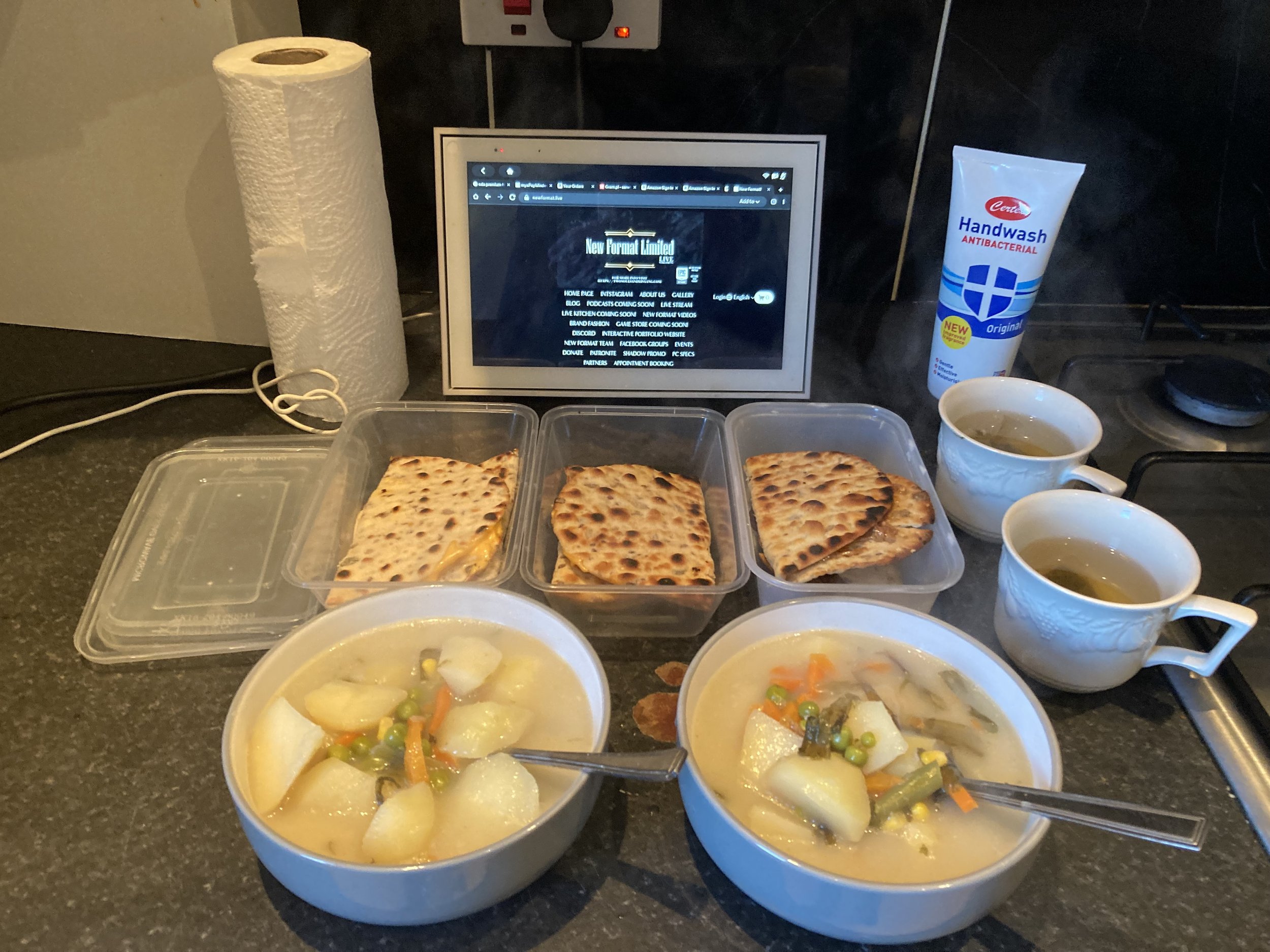 A kitchen counter with containers of flatbread, bowls of vegetable stew, cups of tea, a roll of paper towels, an antibacterial hand wash, and a tablet displaying a website.