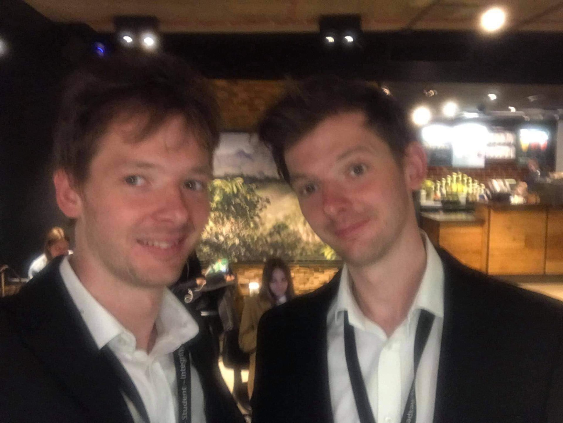 Two young men in suits smiling for a photo in a dimly lit bar or restaurant.