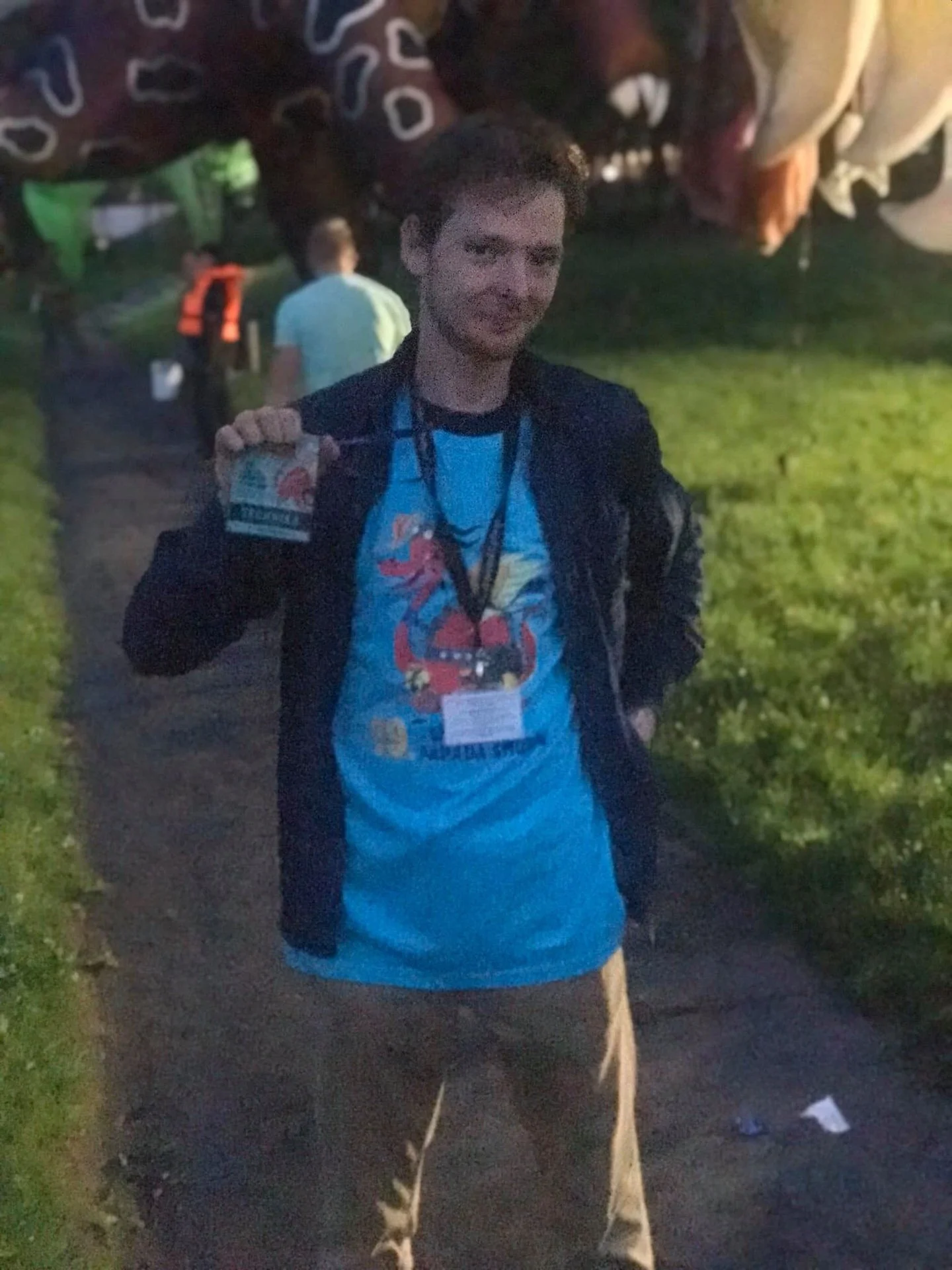 A young man standing outdoors on a dirt path at night holding a ticket or badge in his right hand, wearing a blue t-shirt with a red cartoon character, a dark jacket, and khaki pants, with other people walking in the background.