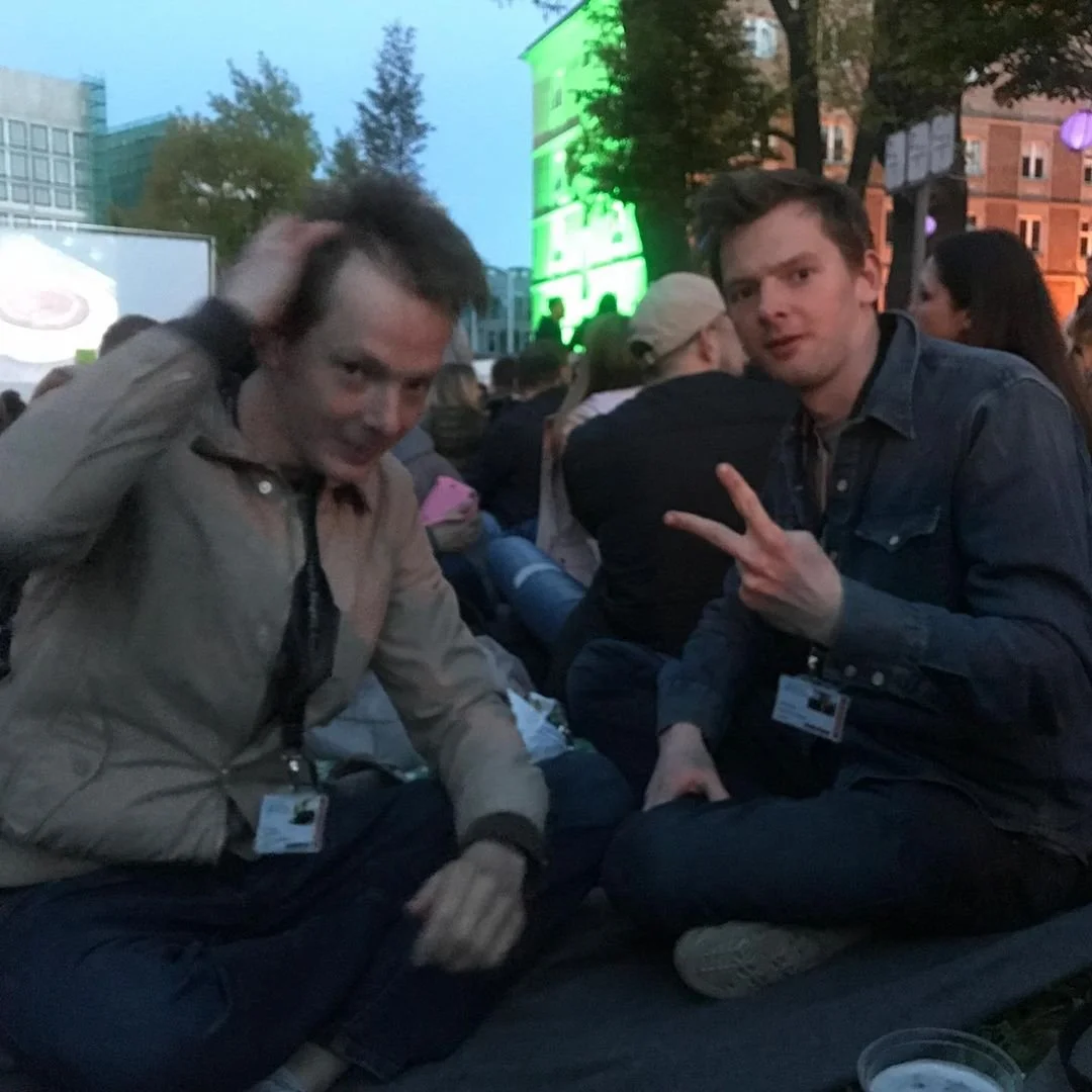 Two young men sitting outdoors at a crowd event during the evening. One man is making a peace sign with his fingers, and the other is scratching his head. There are people and colorful lights in the background.
