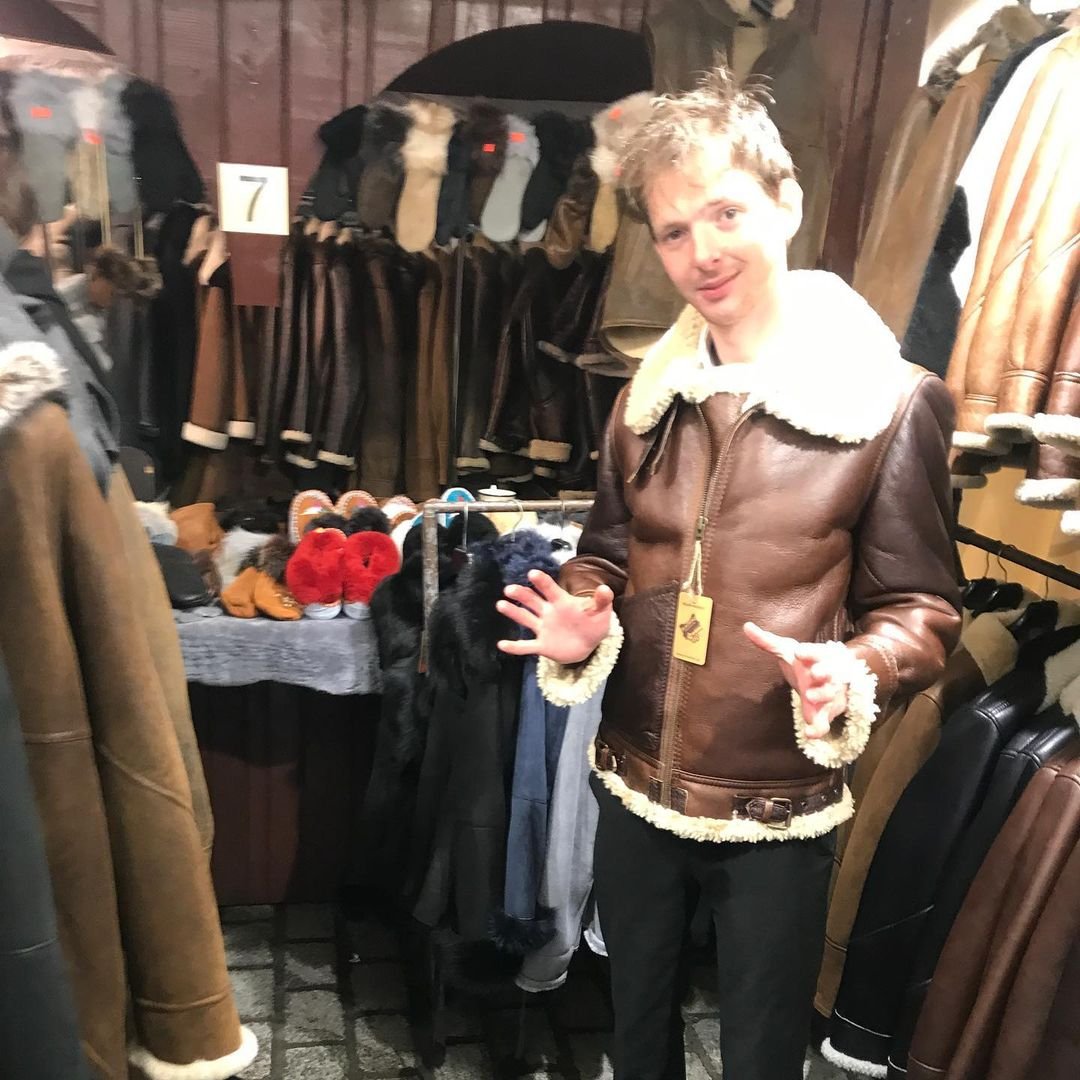 A man in a brown leather jacket with a cream-colored shearling collar stands in a store selling jackets and hats. He is holding up his hand with three fingers and smiling. Behind him are coat racks with various jackets and a table with fuzzy slippers