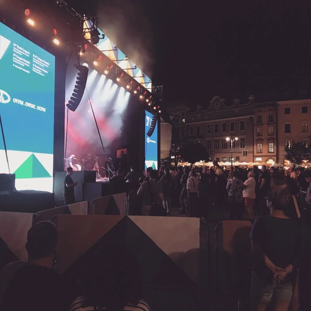 Nighttime outdoor concert with a crowd, stage, and large screens, in an urban setting.