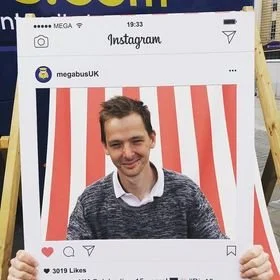 A young man smiling in front of a red and white striped background, appearing inside a large Instagram frame prop.