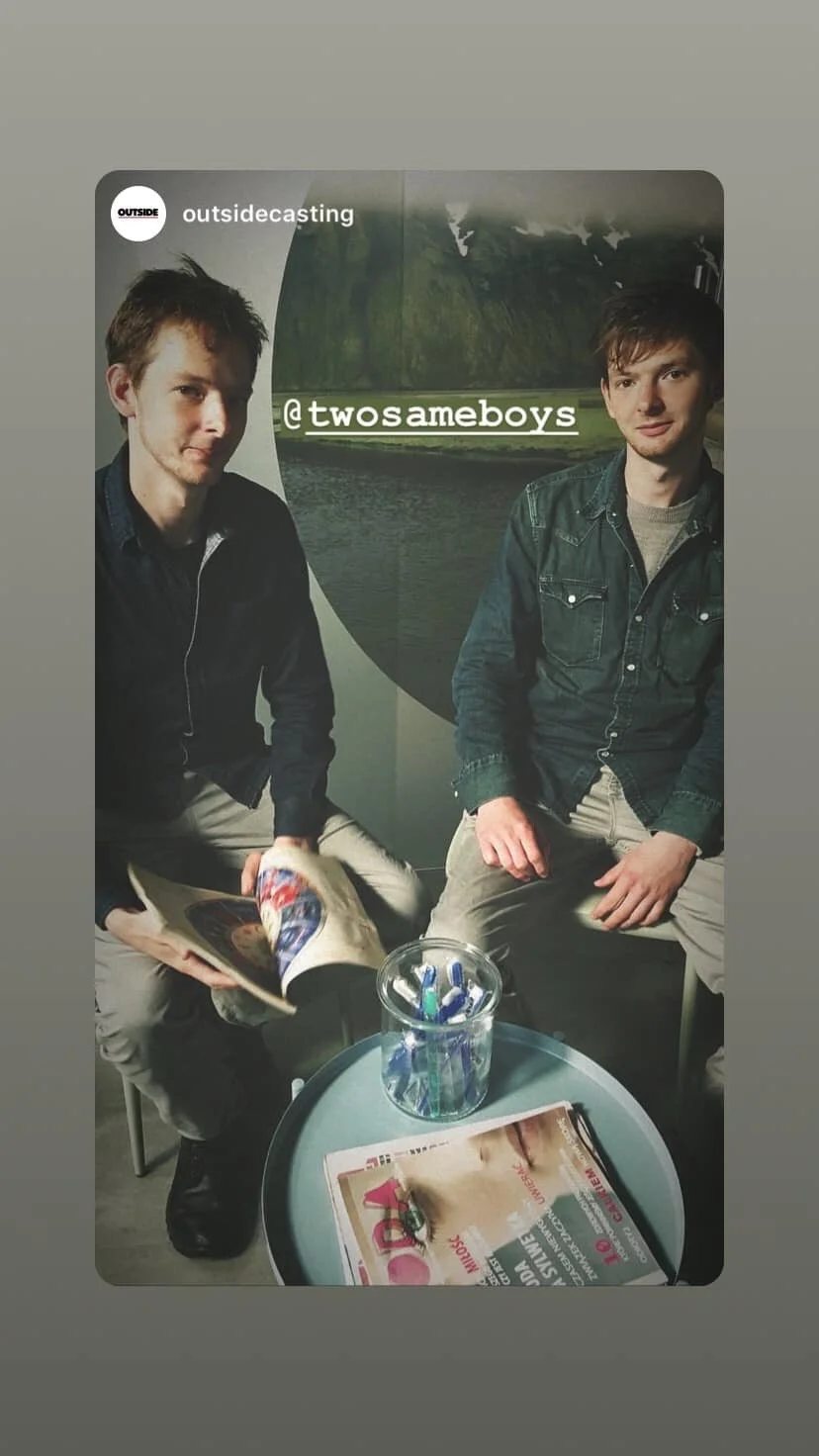 Two young men sitting together in a casual setting, one holding a magazine. There is a small table with a jar of colored pens and newspapers in front of them. The background features a large scenic mural of a landscape with mountains, water, and tree