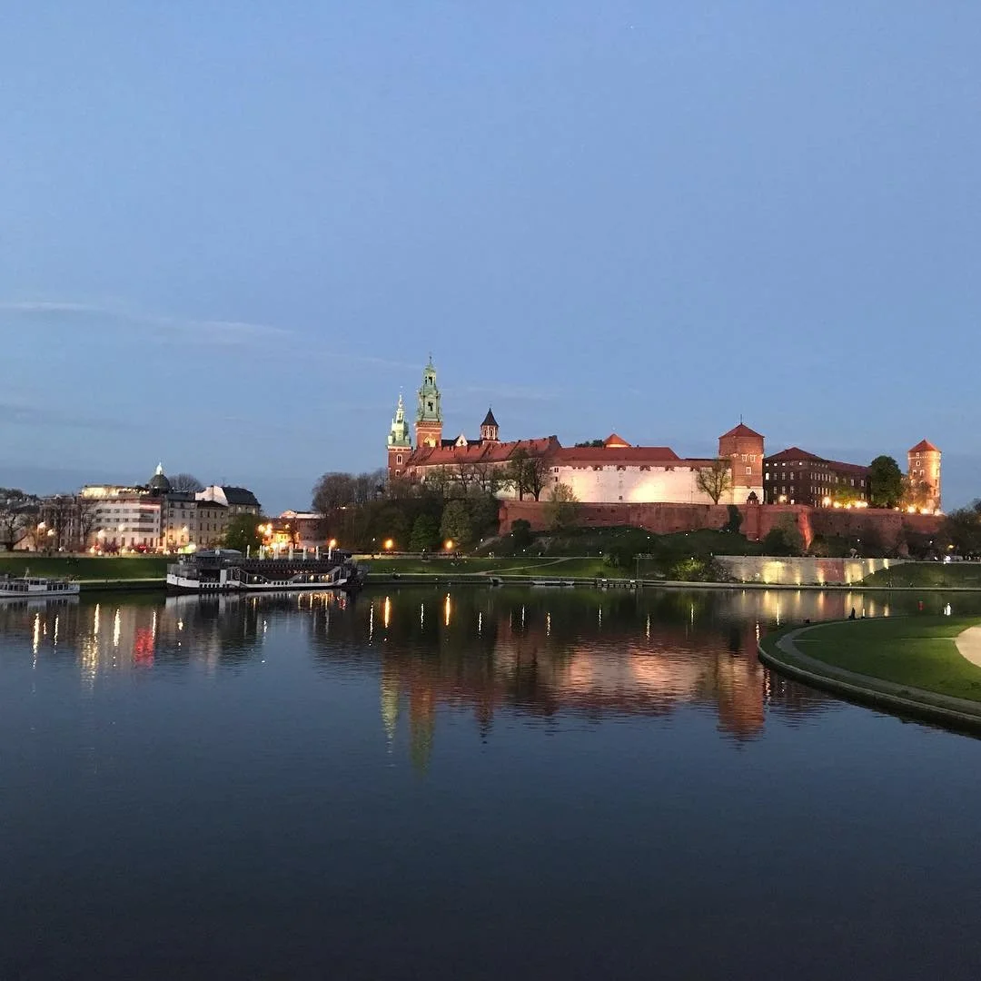 Night view of a historic castle or palace with tall towers and a green-domed tower, reflected in a calm river, with city buildings and trees in the background under a blue sky.
