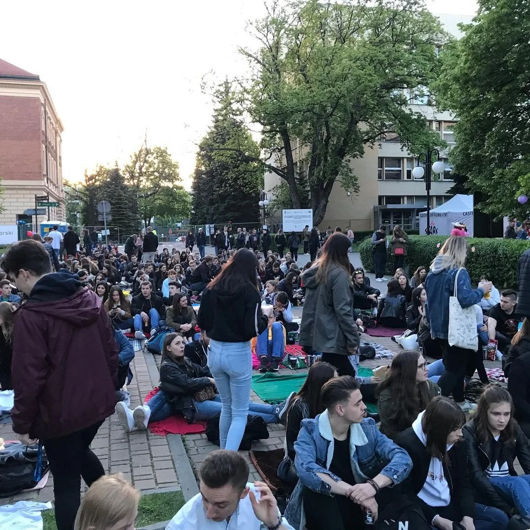 Large group of young people sitting and standing in a park or outdoor area with trees and buildings in the background, gathered for an event or festival.
