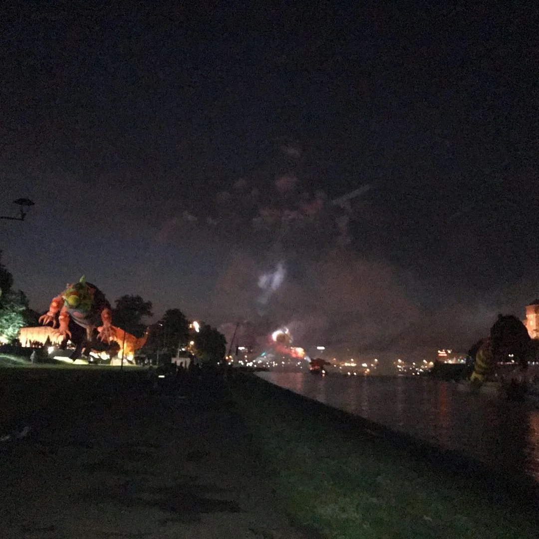 Nighttime scene along a river with fireworks in the sky, large colorful dragon statues on the left side, and city lights reflecting on the water.