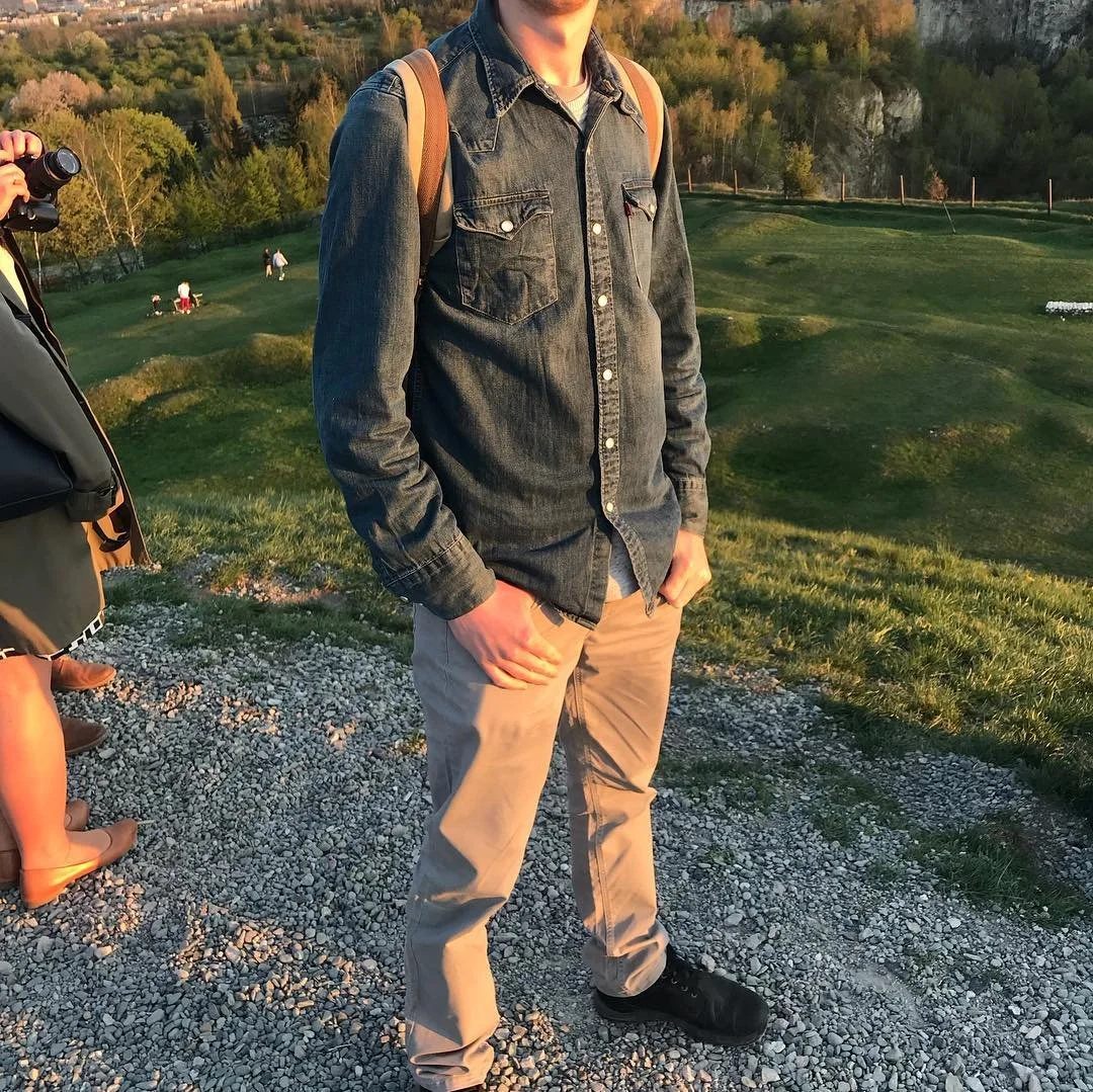 A person standing outdoors on a grassy area during sunset, wearing a denim jacket, beige pants, and black shoes, with a backpack on their shoulder. There are people in the background on a hill and trees in the distance.