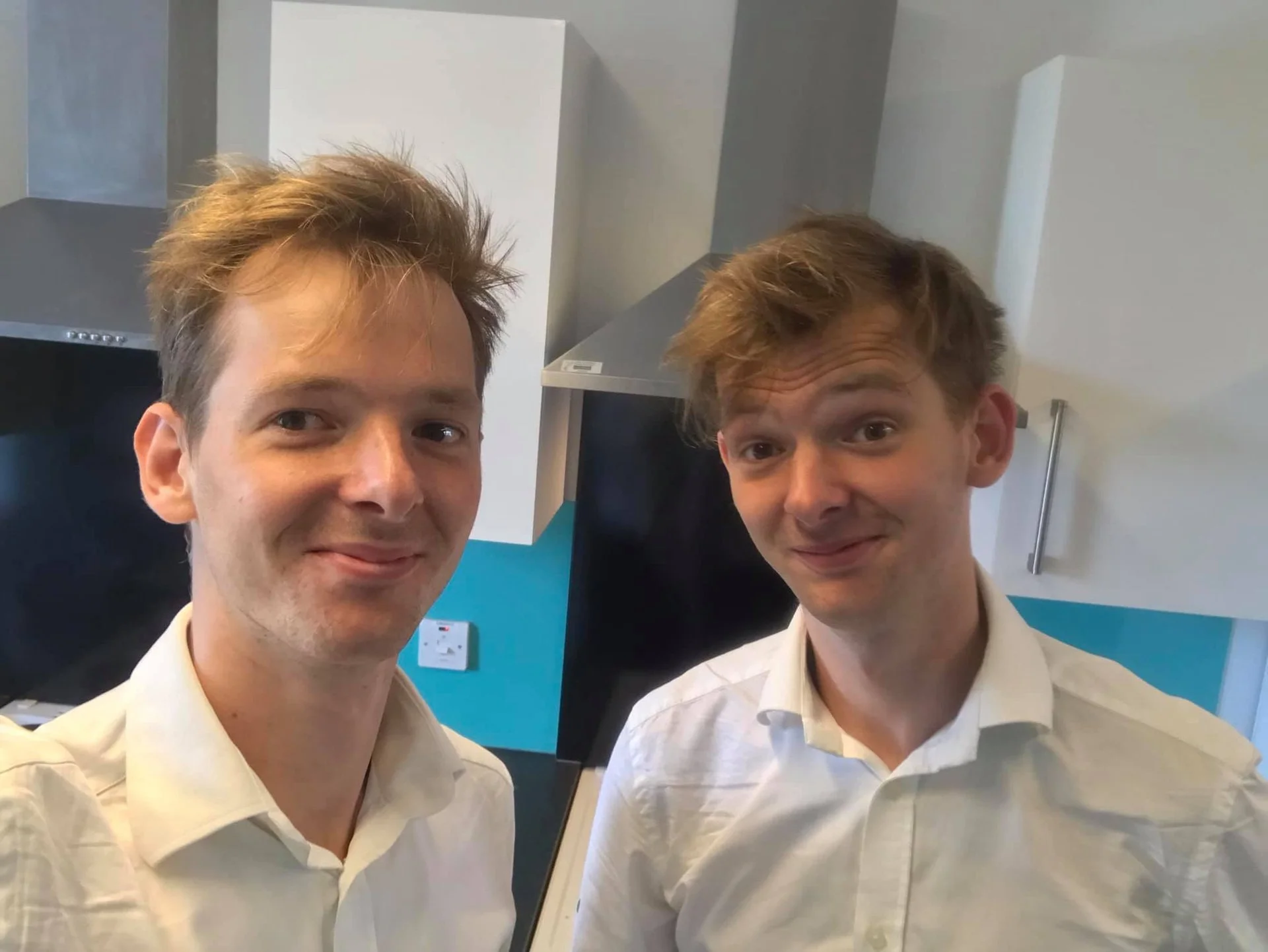 Two men with similar features and light-colored shirts, taking a selfie in a kitchen.