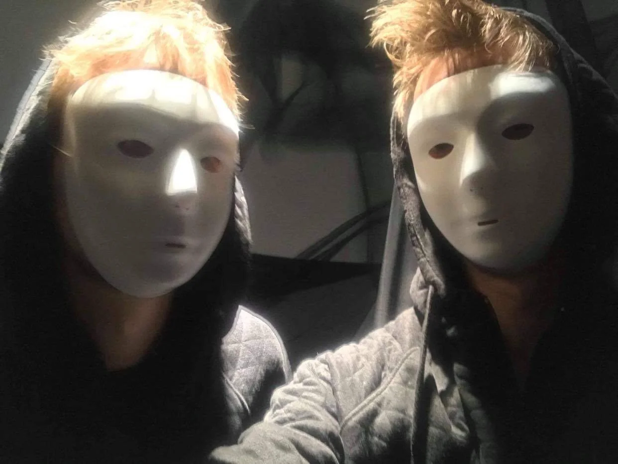 Two individuals wearing white face masks with hollow eyes and lips, dressed in dark clothing and hoodies, in a dimly lit space.