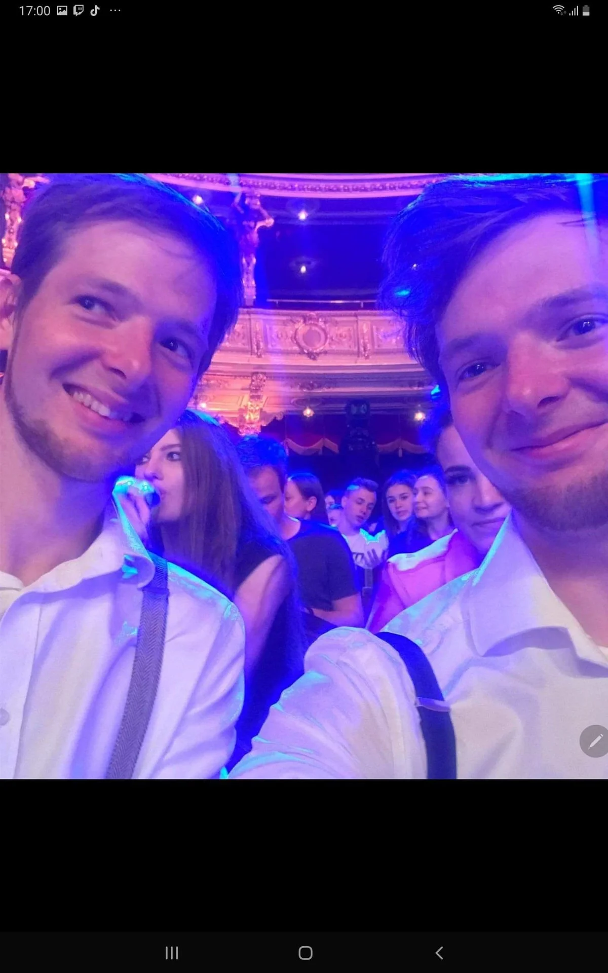 Two young men taking a selfie at a crowded event with ornate architecture and purple lighting.