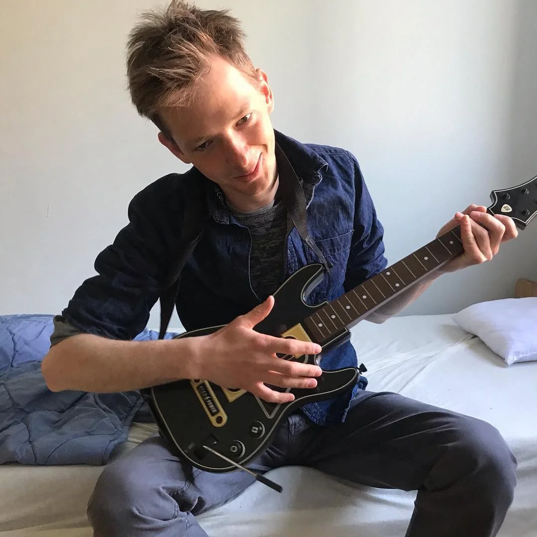A young man with messy blonde hair sitting on a bed, playing a black electric guitar.
