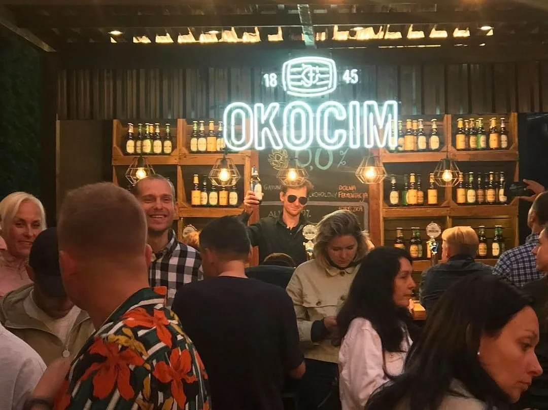 A crowded bar or pub with a wooden backdrop, illuminated signage reading "OKOCIM" in neon lights, bottles of beer on shelves, and people socializing and enjoying drinks, some in casual and tropical patterns.