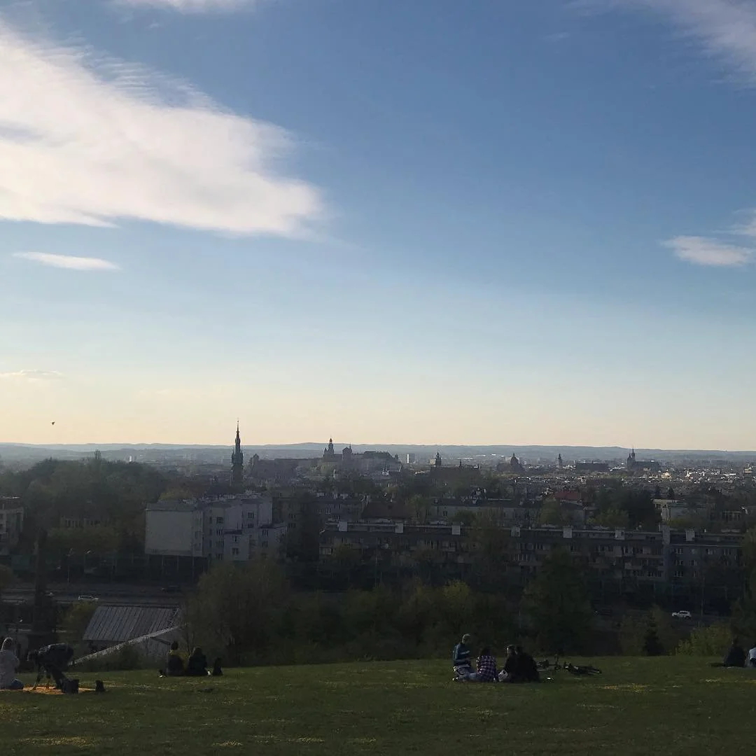 People sitting on a grassy hill overlooking a city skyline with historic buildings and church spires under a partly cloudy sky in the distance.