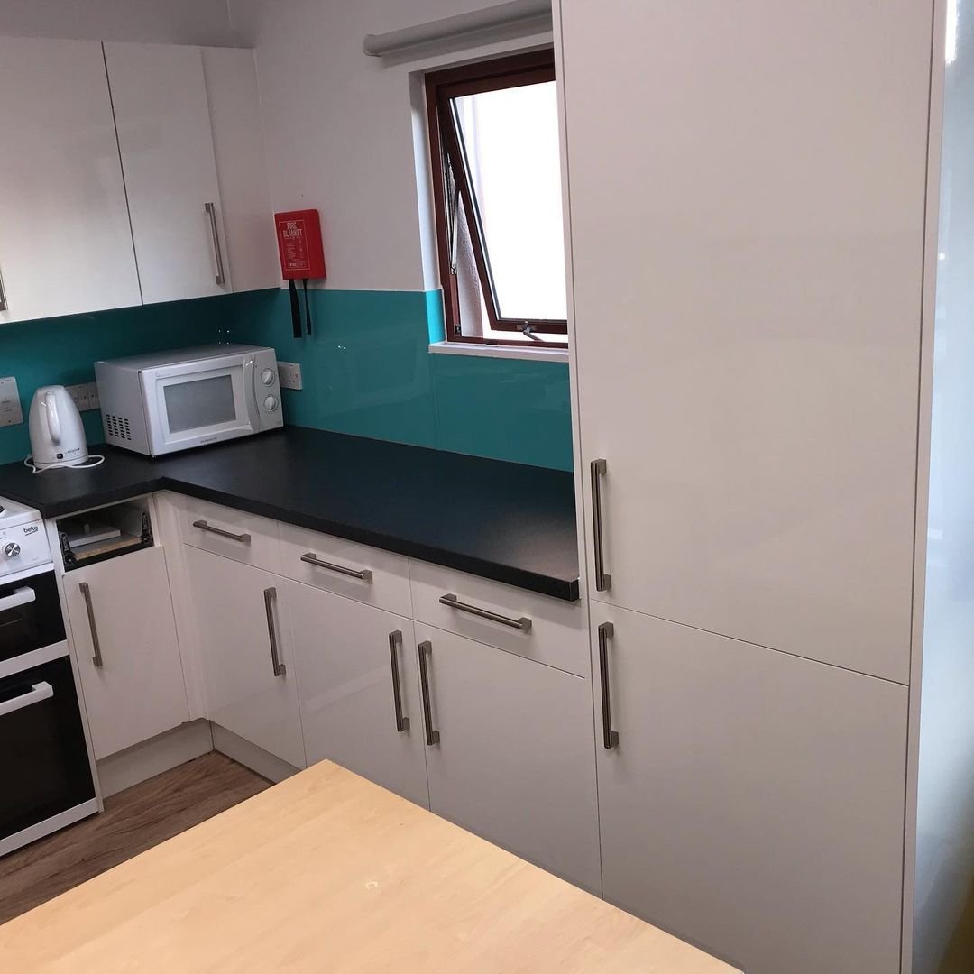 A kitchen corner with white cabinets, a black countertop, a microwave, a kettle, a fire alarm, and a window.