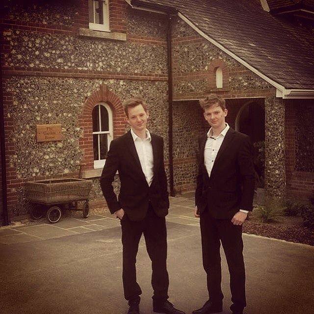 Two young men in formal black suits and white shirts standing outside in front of a brick and stone building.