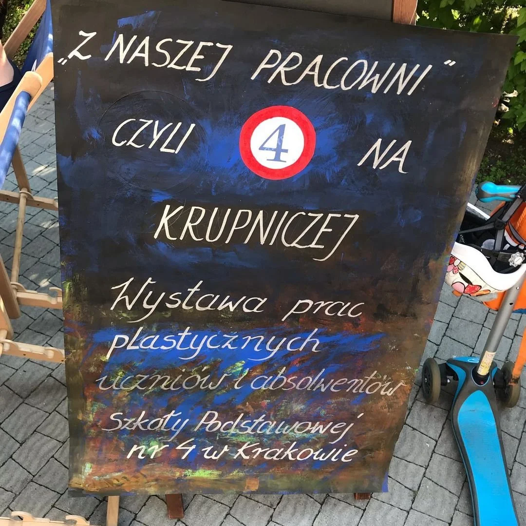 Blackboard sign with educational-themed colorful, artistic background, featuring Polish text about a creative exhibition at a primary school in Krakow, with a large red and white number 4 in the center.