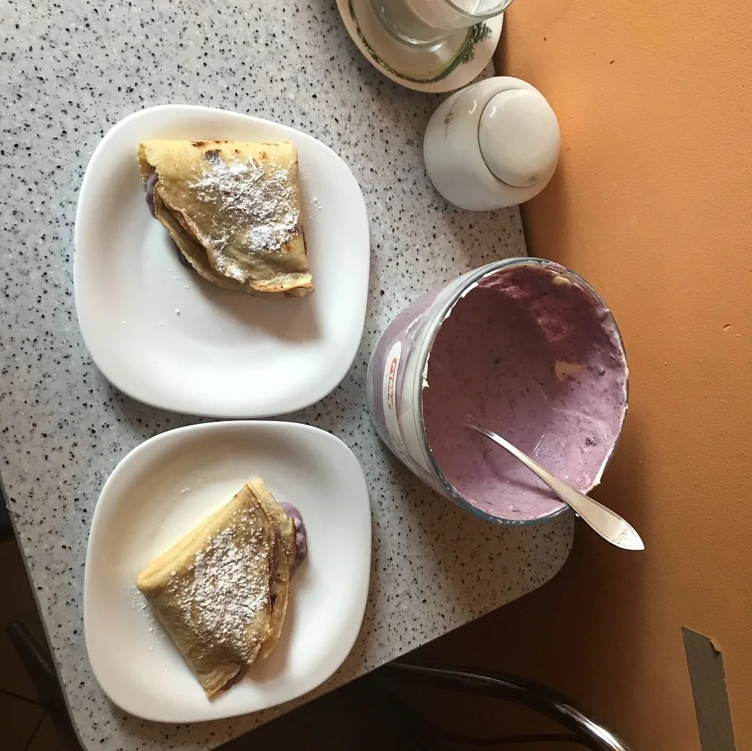 Two plates of pie, one blueberry and one cherry, with powdered sugar, a glass of milk, a jar of pink berry smoothie, a salt shaker, and a pepper shaker on a speckled countertop.