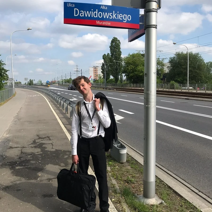Young man with a backpack and carrying a black duffel bag standing on a sidewalk next to a street with train tracks, under a street sign that reads 'Dawidowskiego' on a partly cloudy day.