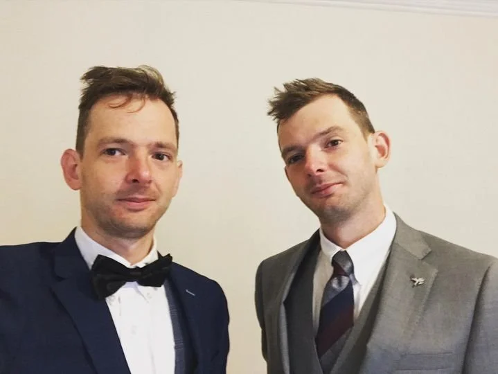 Two men in formal suits posing for a photo against a plain wall.