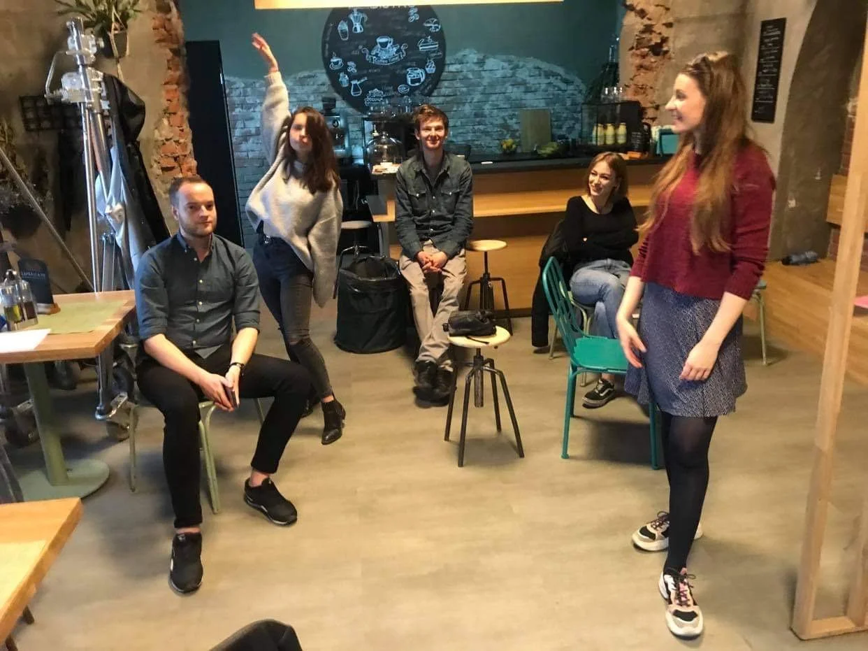 Five people in a cozy cafe or restaurant, with two sitting and three standing or moving, engaged in conversation or interaction.