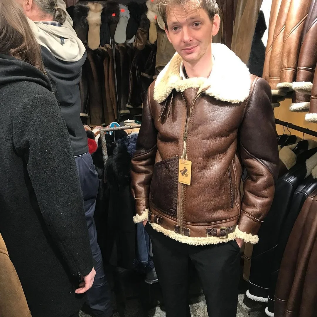 A young man trying on a brown leather aviator jacket with a shearling collar in a store. The store has other jackets and coats hanging around.