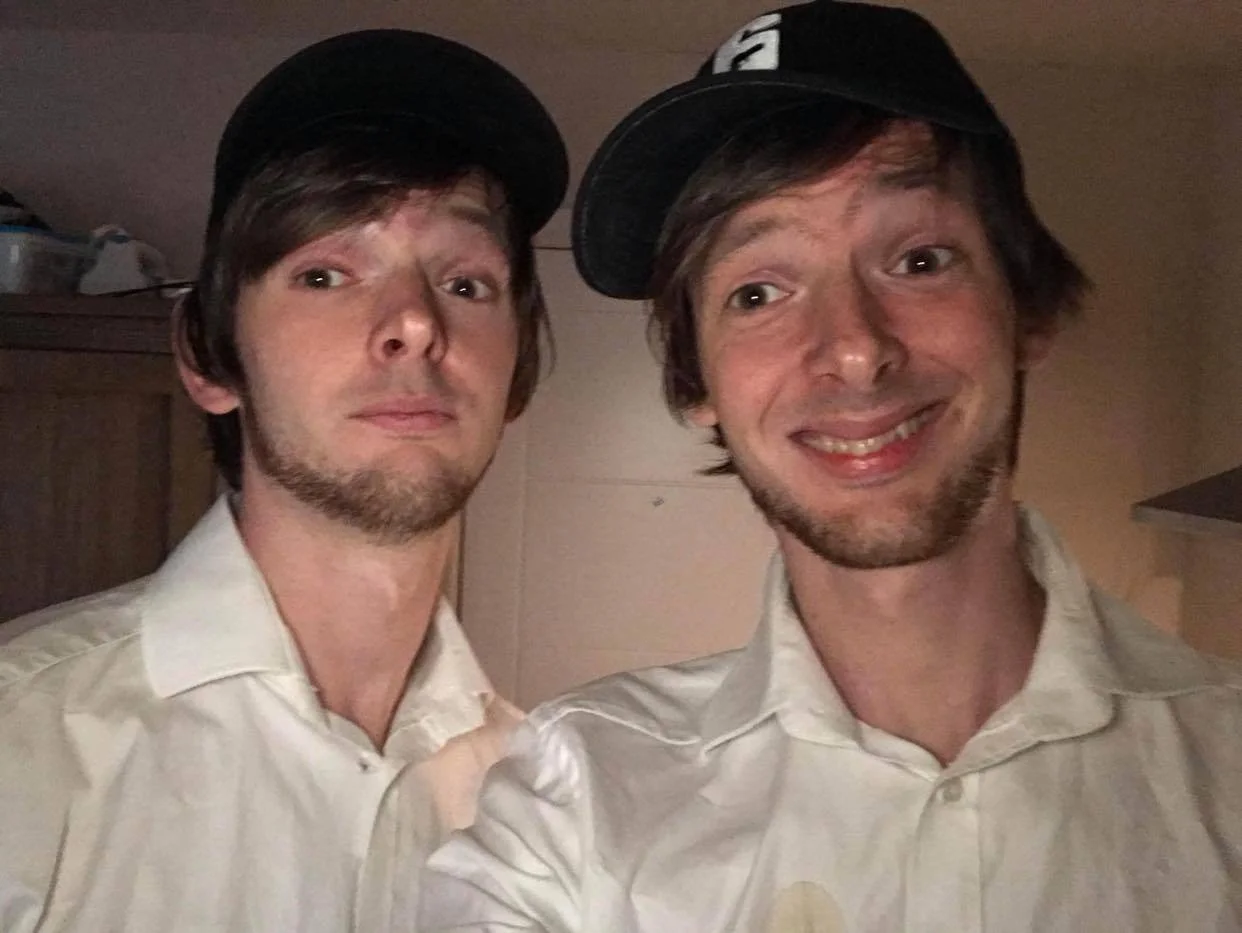Two young men with light skin and brown hair taking a selfie indoors. One is making a serious face, and the other is smiling. Both are wearing white shirts and black caps.