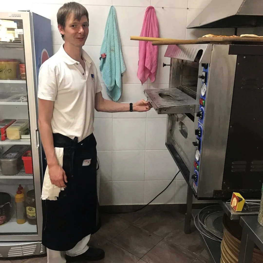 A man stands in a kitchen near a pizza oven, holding the oven door open. He is wearing a white polo shirt and an apron. To his left, there is a freezer with various items inside, and in the background, two towels hang on the wall.