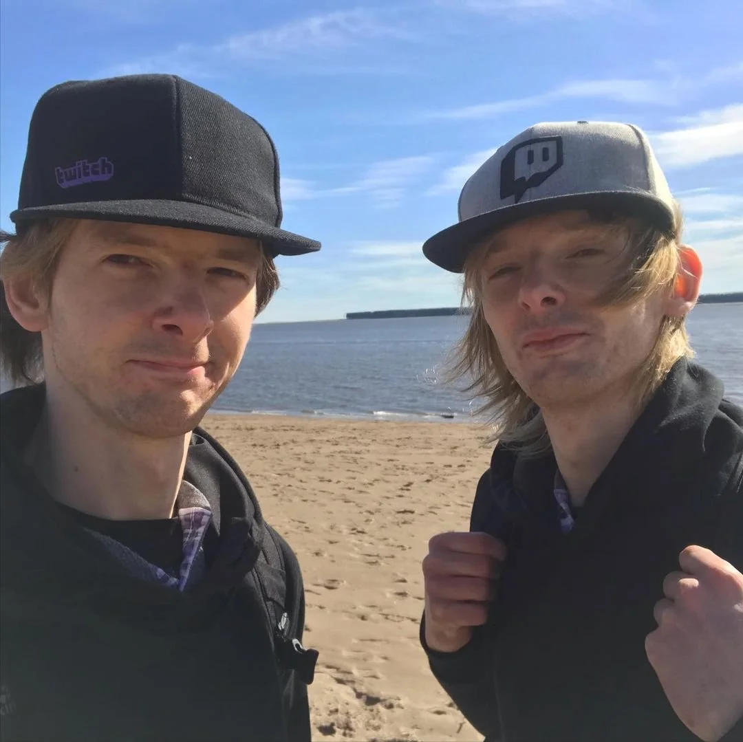 Two young men with long hair and light skin taking a selfie on a sandy beach by a body of water under a blue sky. Both are wearing baseball caps with the Twitch logo and backpacks.