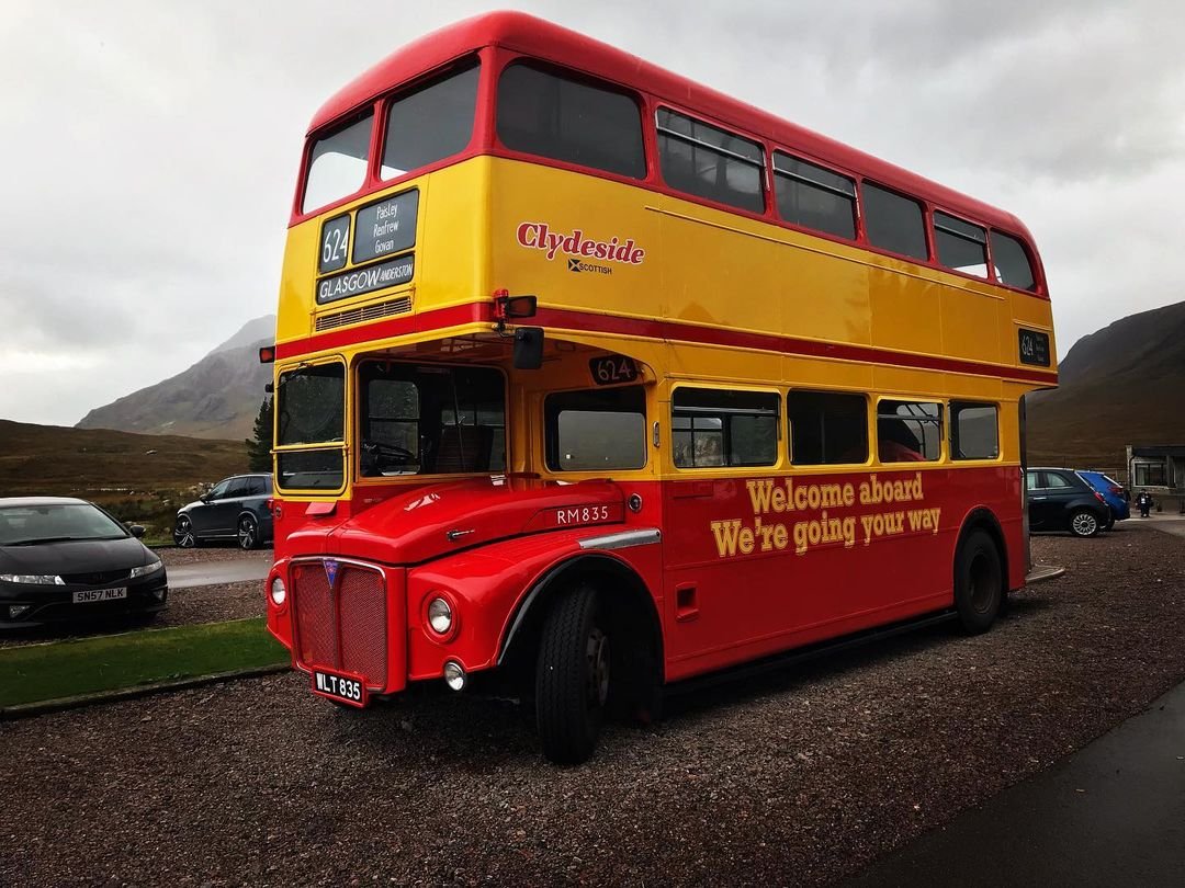A double-decker bus painted in red and yellow with the words "Welcome aboard We're going your way" on the side, parked outdoors with mountains in the background and several cars nearby.
