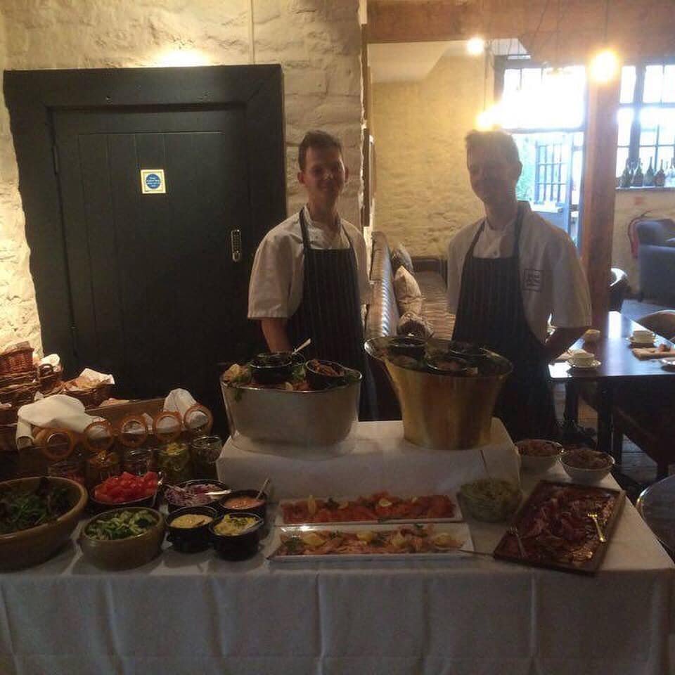 Two chefs standing behind a table with salads, sauces, and food trays in a restaurant.