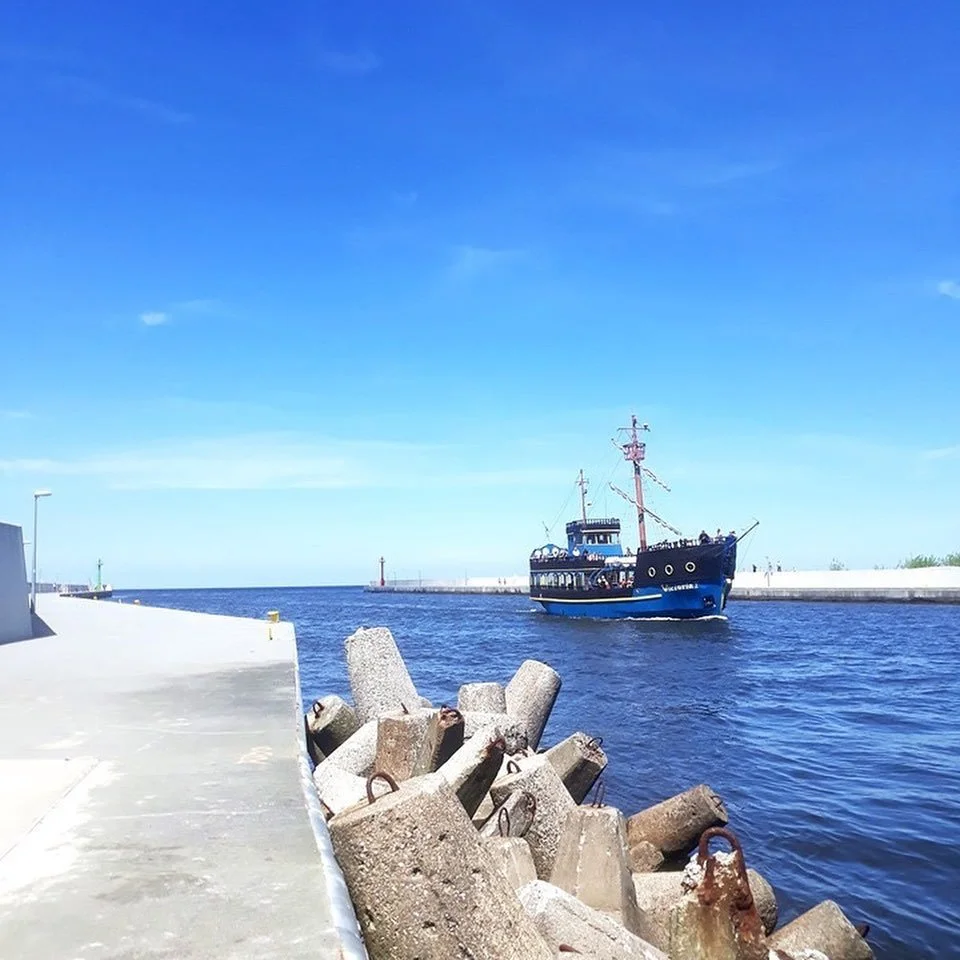 A blue boat sailing in a harbor with a concrete wall and large concrete blocks along the shore, and a lighthouse in the distance.