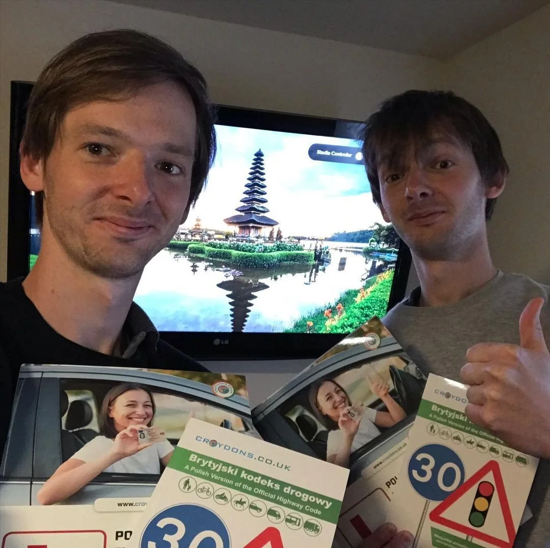 Two men taking a selfie indoors in front of a television screen displaying a traditional Asian temple with water reflecting the structure. They are holding flyers and smiling, with one giving a thumbs-up.