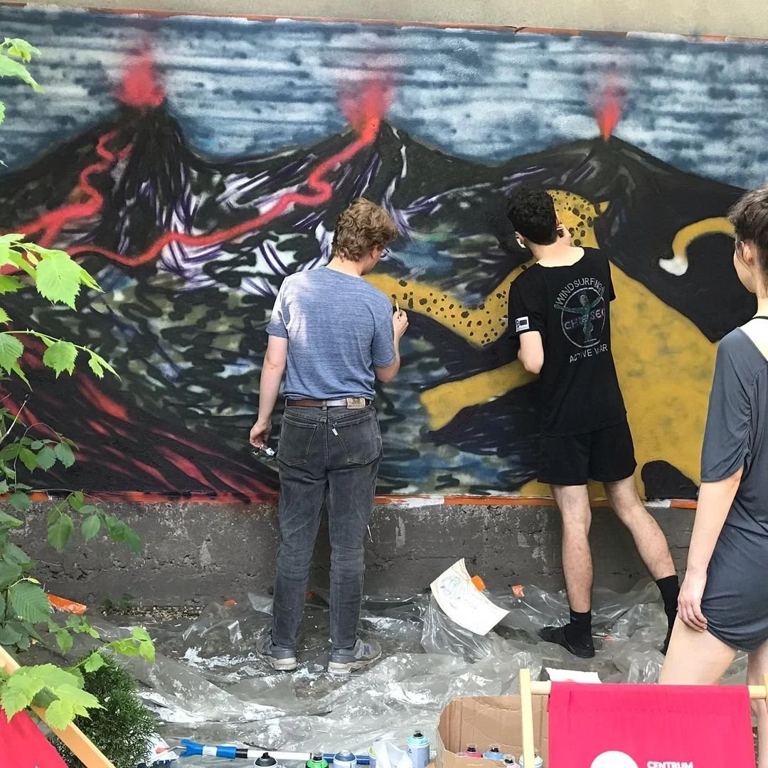 Four people are painting a mural of erupting volcanoes on a wall. The mural depicts two volcanoes with flowing lava and smoke. The artists are standing on a plastic sheet on the ground, and there are spray paint cans and art supplies nearby.