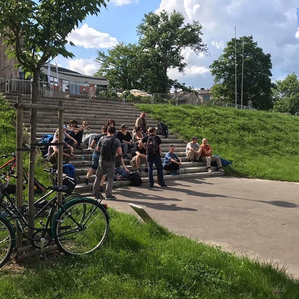 A group of people is sitting and standing on a set of outdoor stairs in a park on a sunny day, with trees and a grassy hill behind them. Some bicycles are parked nearby.