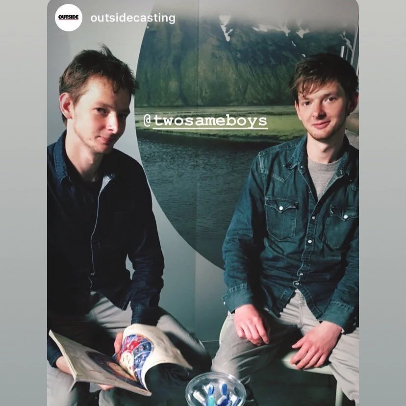 Two young men sitting indoors with a mountain landscape photograph on the wall behind them. One is holding an open magazine, the other has his hands on his lap. Text overlay says '@twosexmeboys' and 'outsidecasting'.