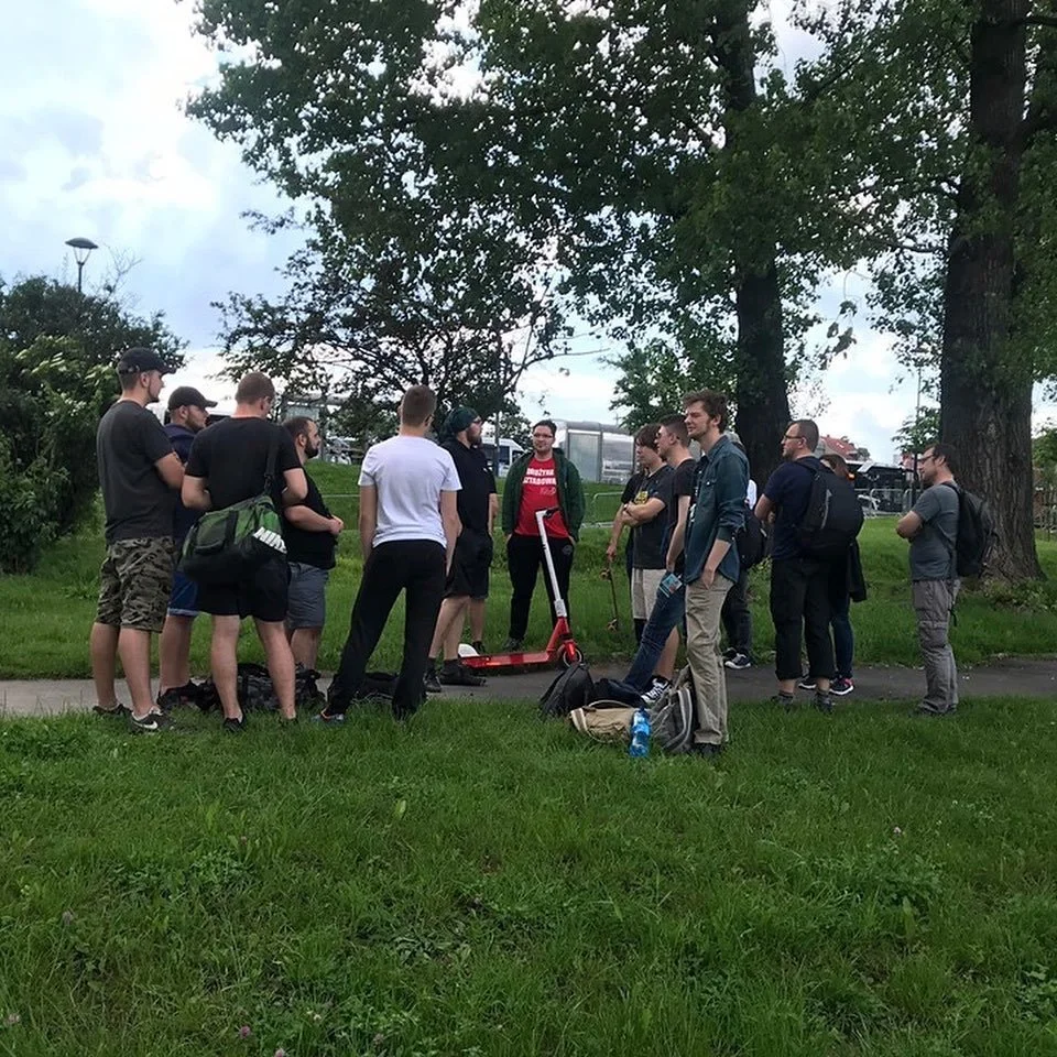 A group of people gathered outdoors on a sidewalk in a park-like setting, some carrying backpacks and personal items, with trees and cloudy sky in the background.