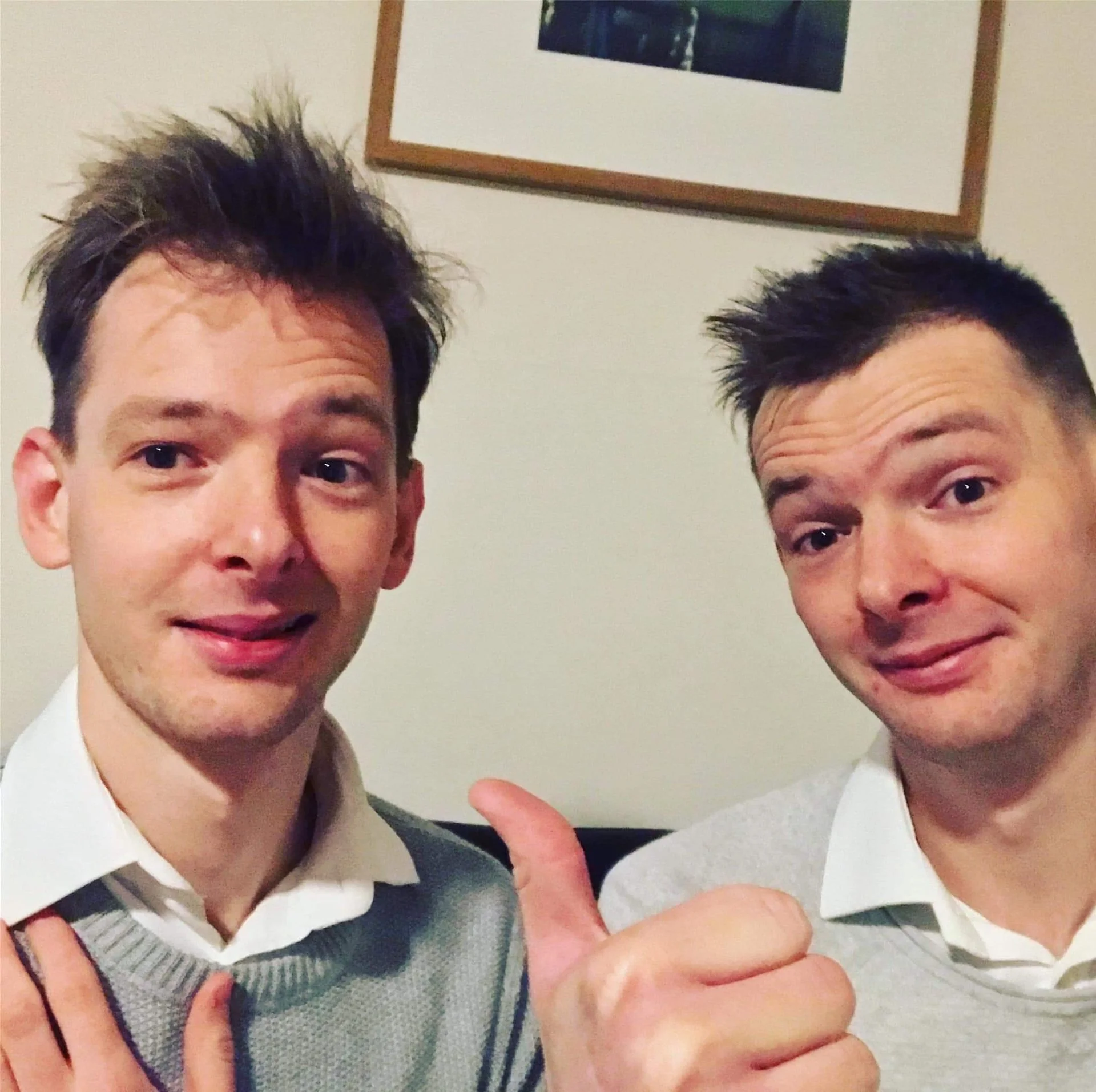 Two young men taking a selfie indoors with light-colored walls and a framed picture hanging behind them. Both are smiling; the man on the left has messy brown hair and is wearing a white shirt with a gray sweater, while the man on the right has short