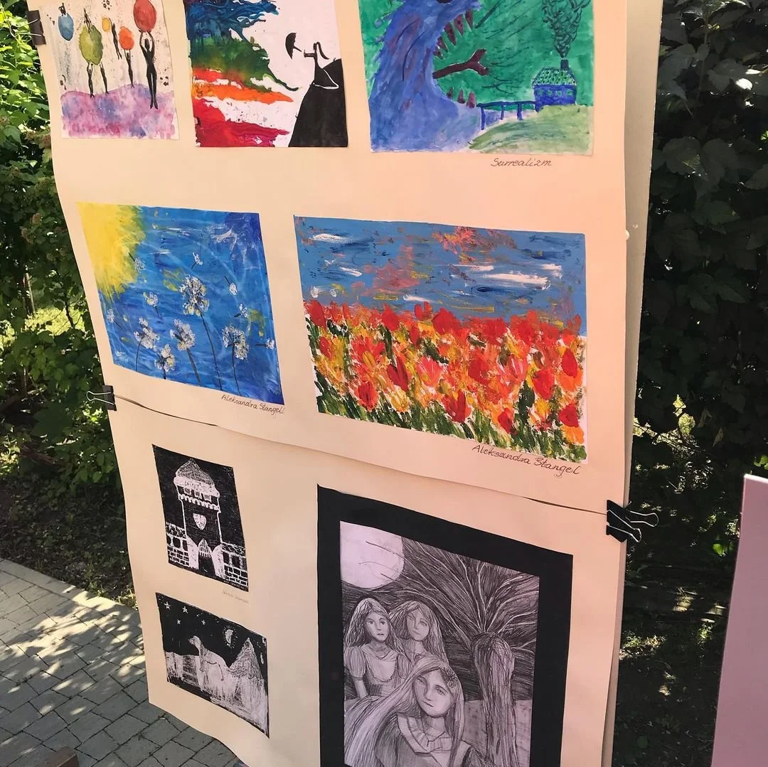 Display of various children's art pieces including colorful paintings of flowers, landscapes, and abstract scenes, as well as black and white drawings of buildings and people, mounted on a board outdoors.