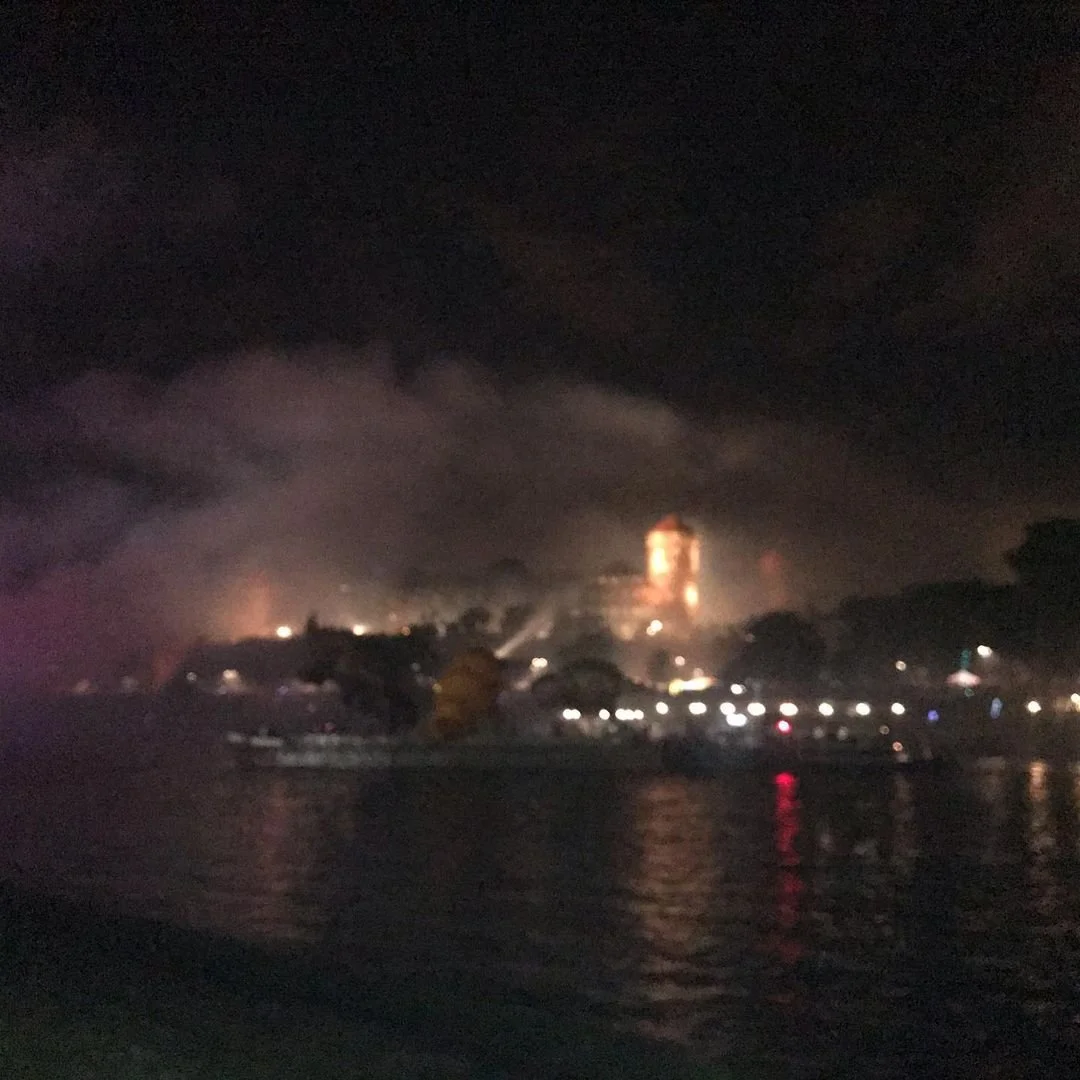 Nighttime cityscape with a large building illuminated, viewed across a body of water with reflections of lights, and dark clouds above.