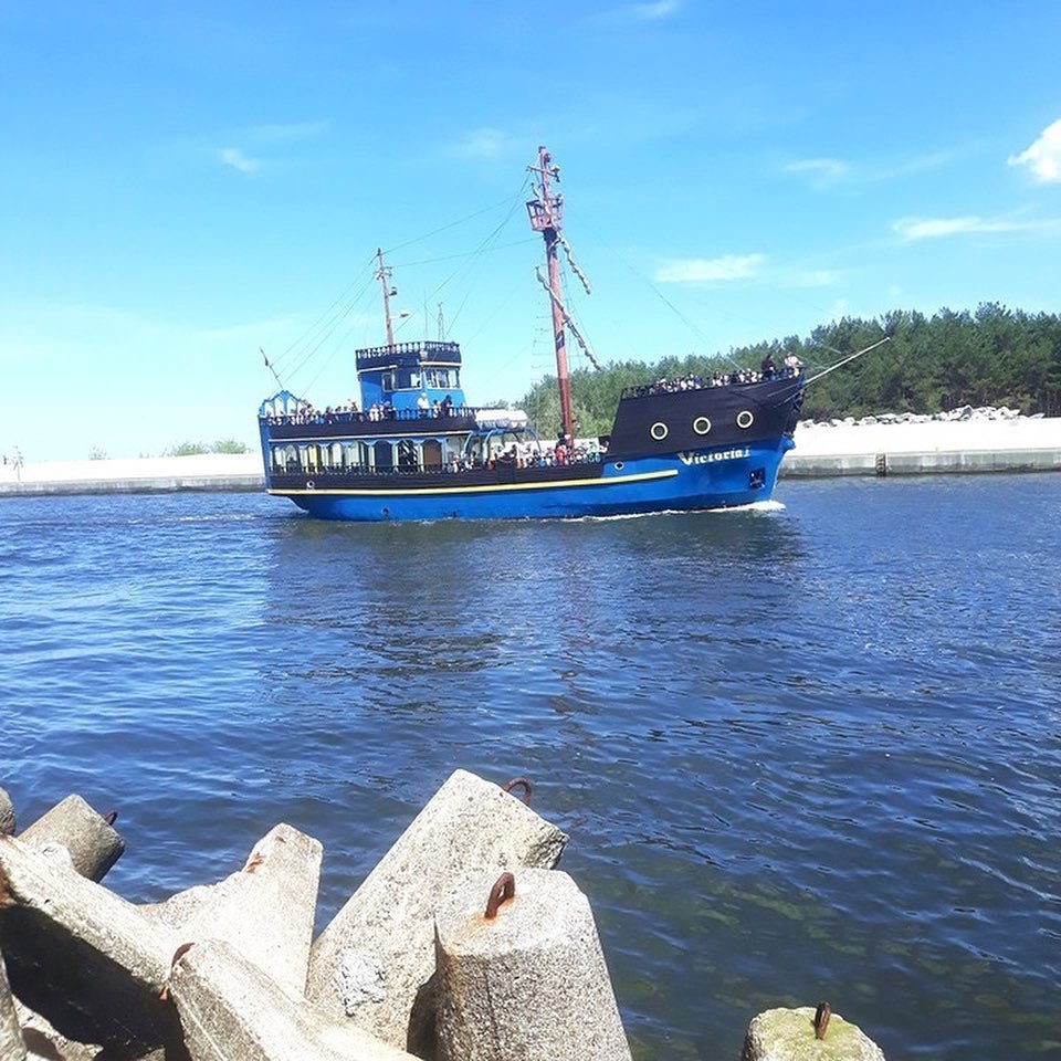 A blue and black boat named Victoria sailing on a calm body of water near a shoreline with trees and rocks.