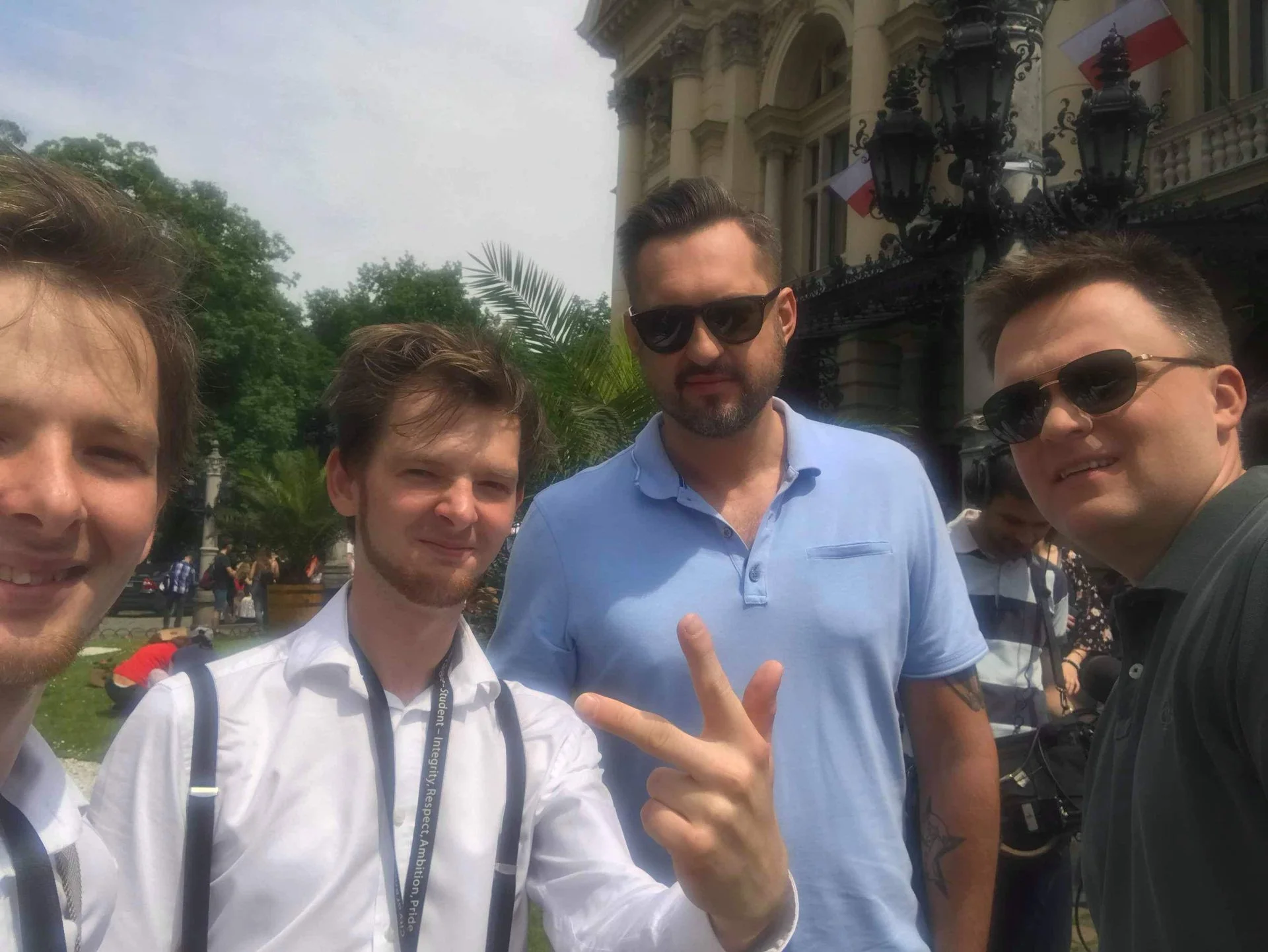 Four men taking a selfie outdoors in front of a historic building with ornate black lamps and flags. Some are wearing sunglasses, one is flashing a peace sign.