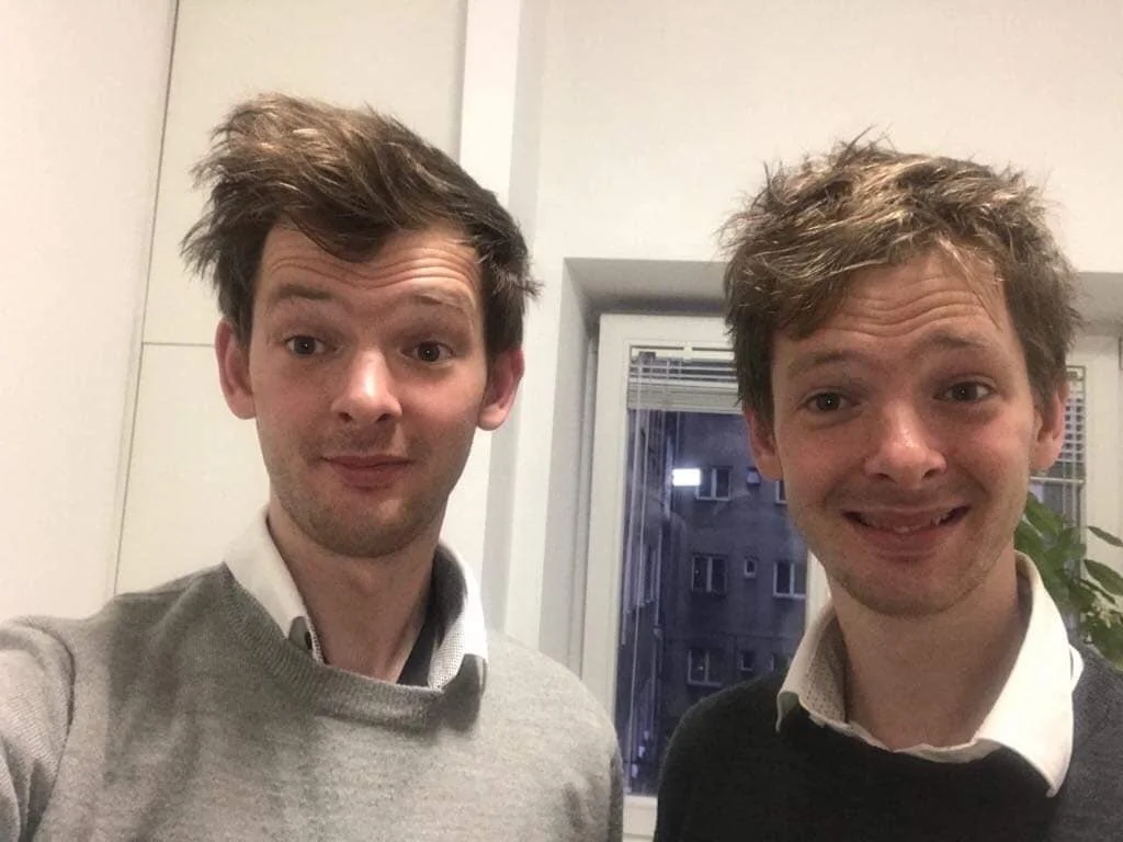 Two young men with messy hair taking a selfie indoors near a window.