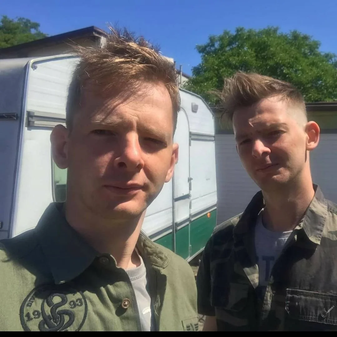 Two young men with short styled hair taking a selfie outdoors with a camper trailer behind them and green trees in the background on a sunny day.