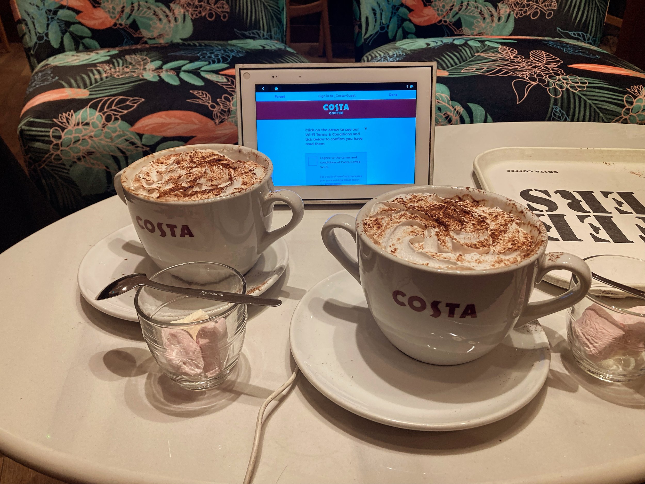 Two large cups of coffee with whipped cream and cocoa powder on top, placed on saucers at a table with a tablet displaying Costa Coffee, small glass of cream, and a tray with a menu or placemat.