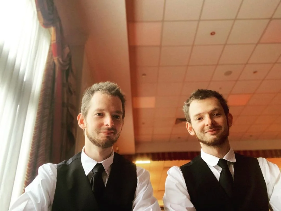 Two young men dressed in white shirts, black vests, and black ties sitting indoors in a room with a wooden ceiling and curtains, smiling at the camera.