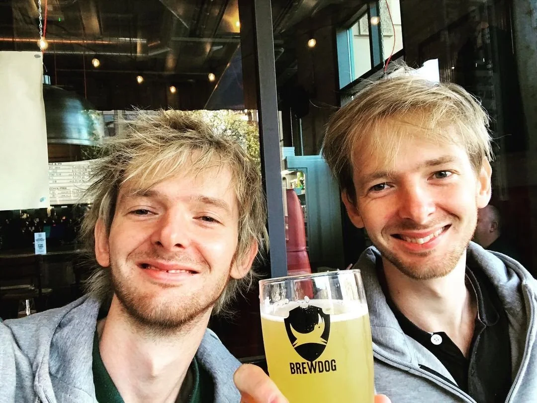 Two young men smiling at a camera while sitting in a restaurant or bar, with one holding a glass of beer that has a Brewdog logo on it.